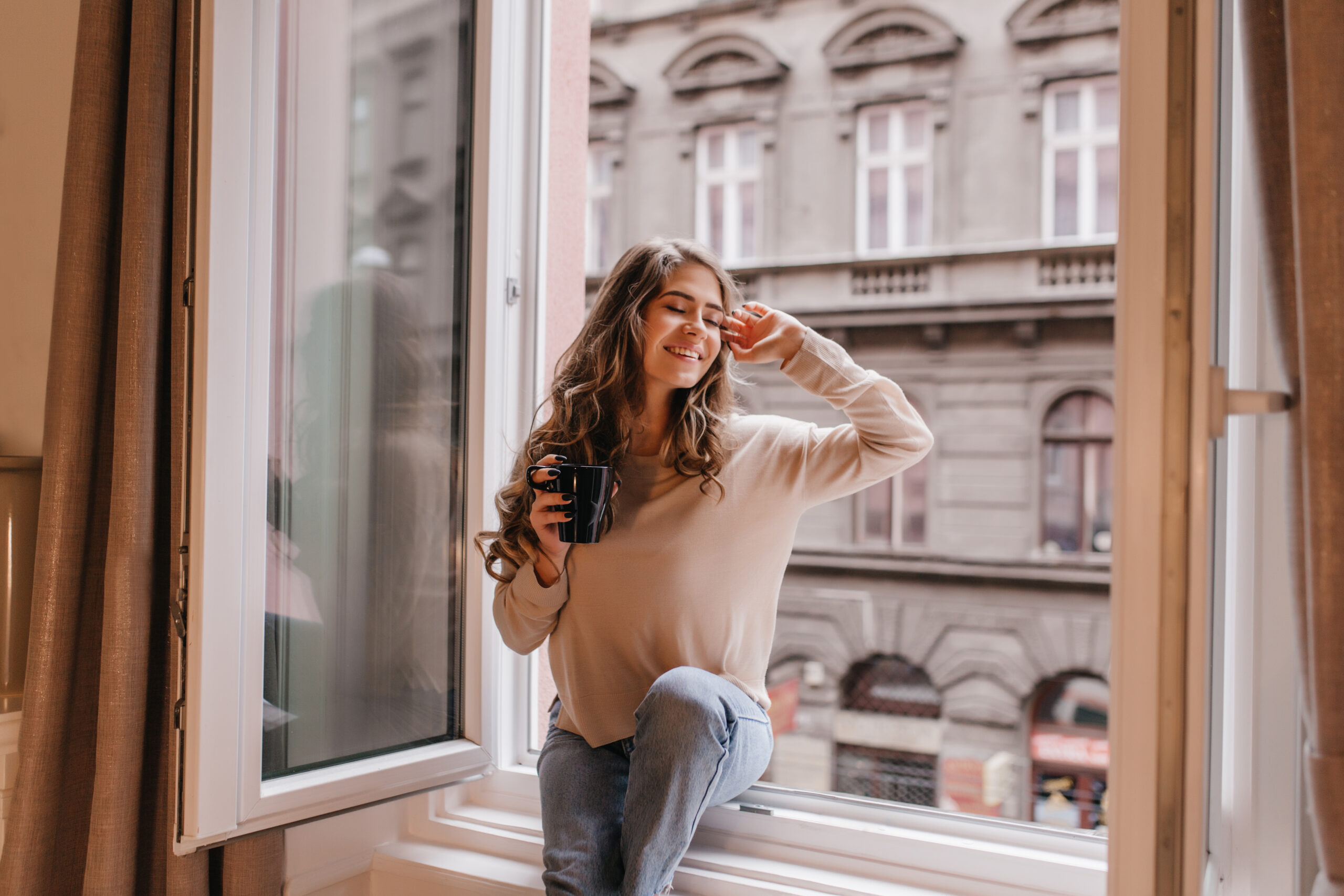 Refined caucasian girl in trendy shirt enjoying city views from window. Attractive curly woman chilling at home, sitting on sill with smile..